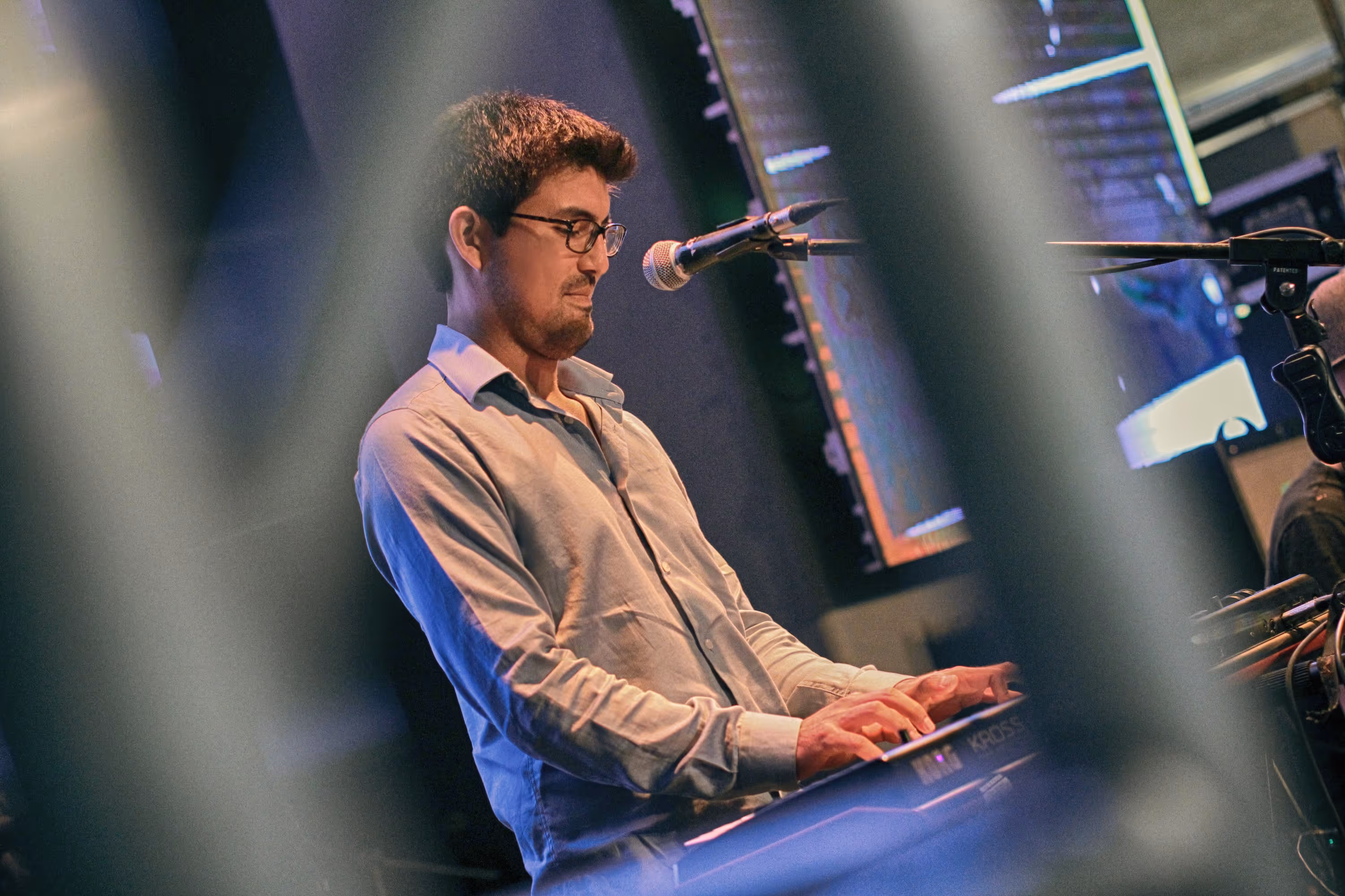 Primer plano de un músico concentrado con lentes tocando un teclado sintetizador en el escenario, fotografiado a través de un desenfoque en primer plano.