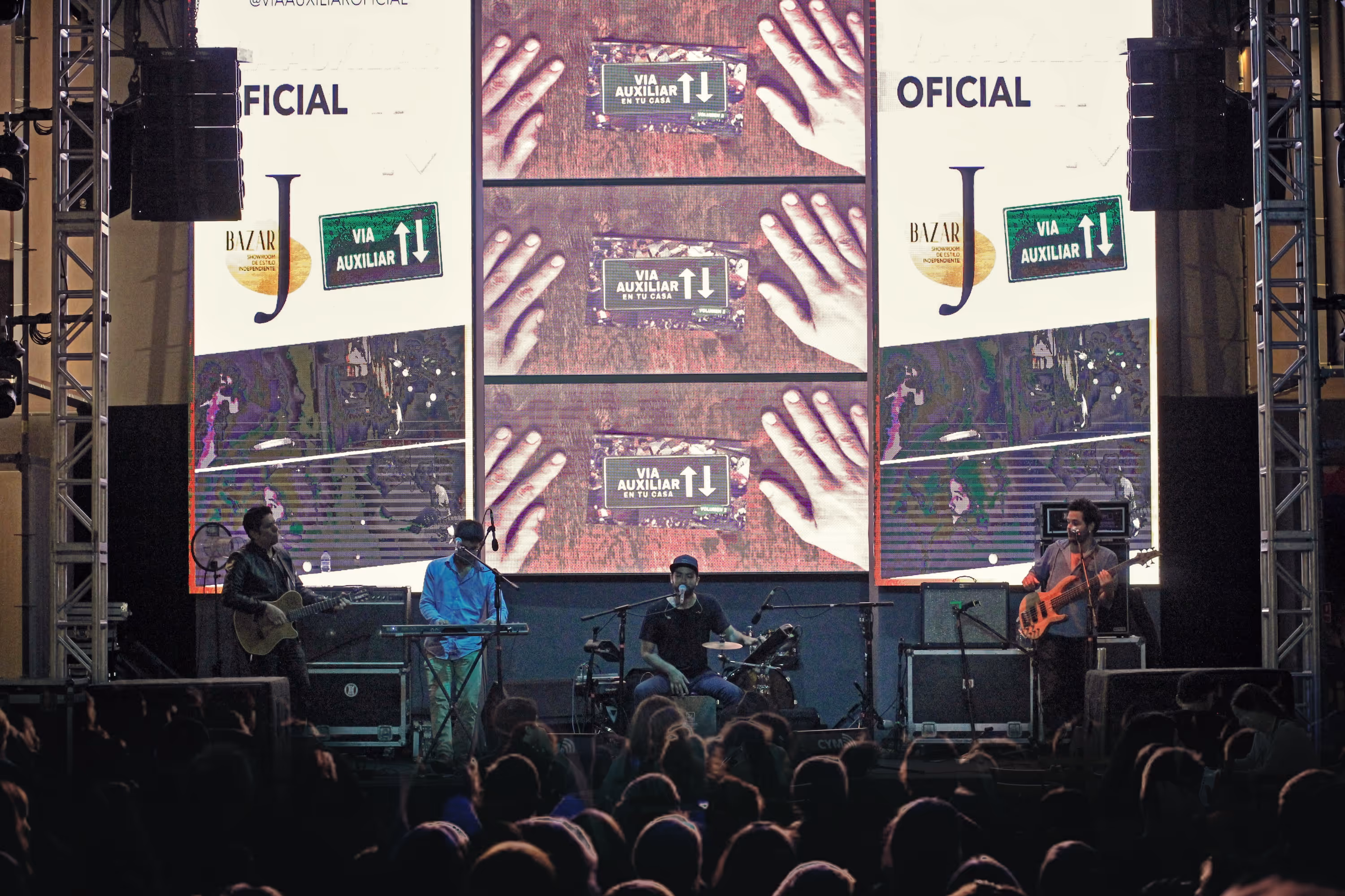 Una banda de cuatro integrantes toca en un gran escenario con pantallas LED masivas detrás mostrando el logo de "Vía Auxiliar", frente a un público numeroso.