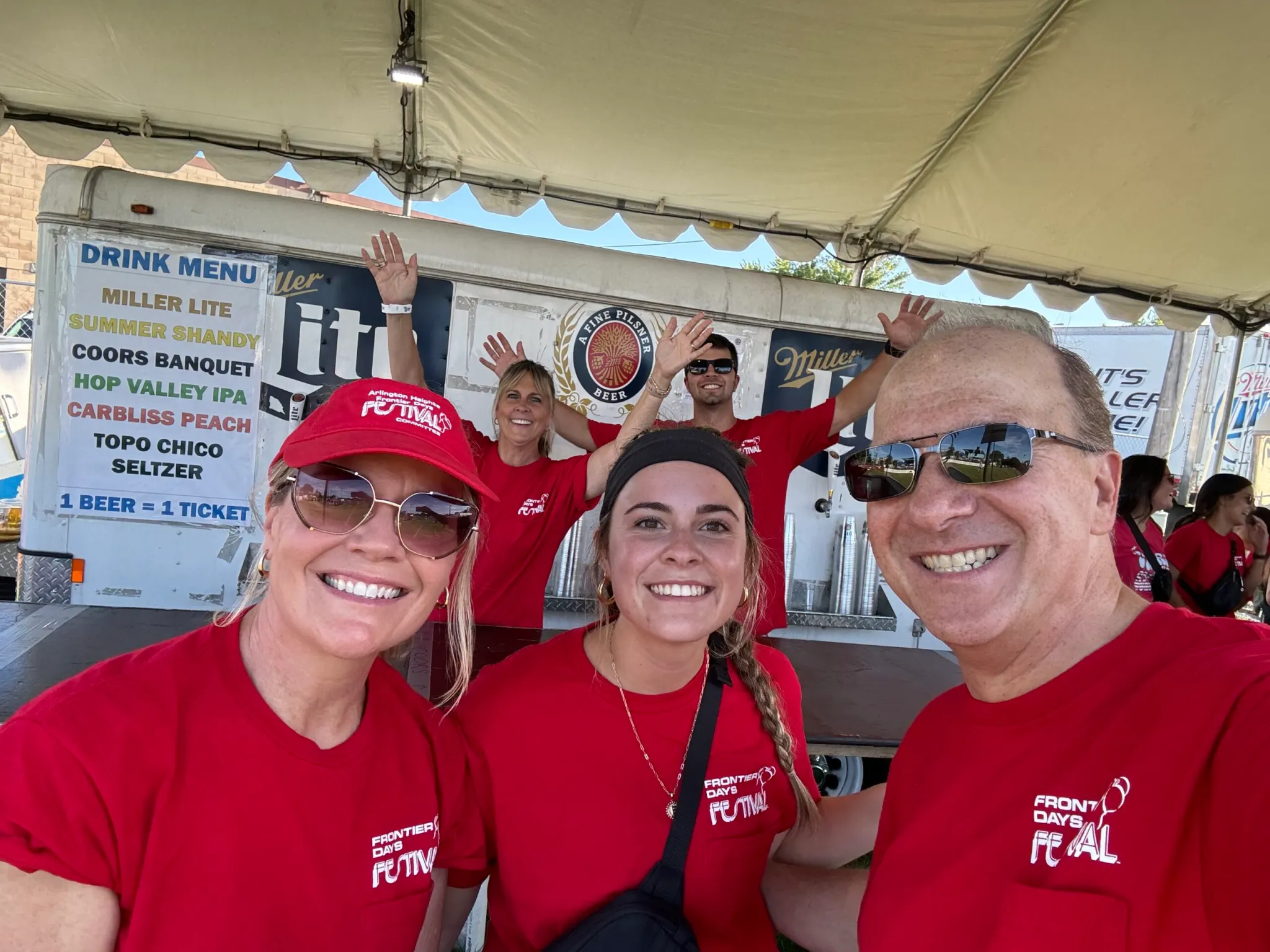 Group of smiling festival volunteers in red Frontier Days Festival shirts posing under a tent by a drink menu board.