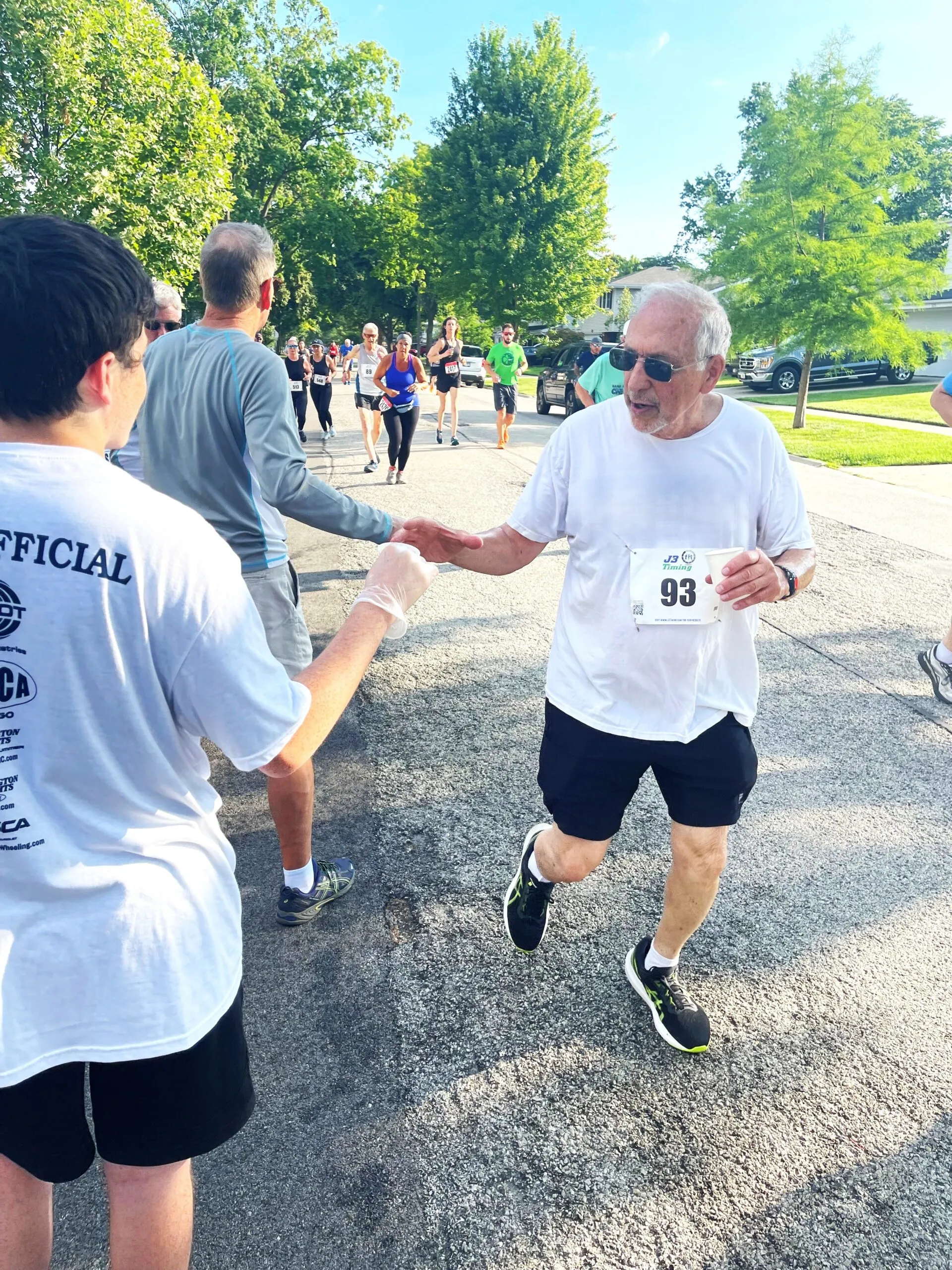 Older male runner with bib 93 fist bumps volunteer during a neighborhood road race on a sunny day.