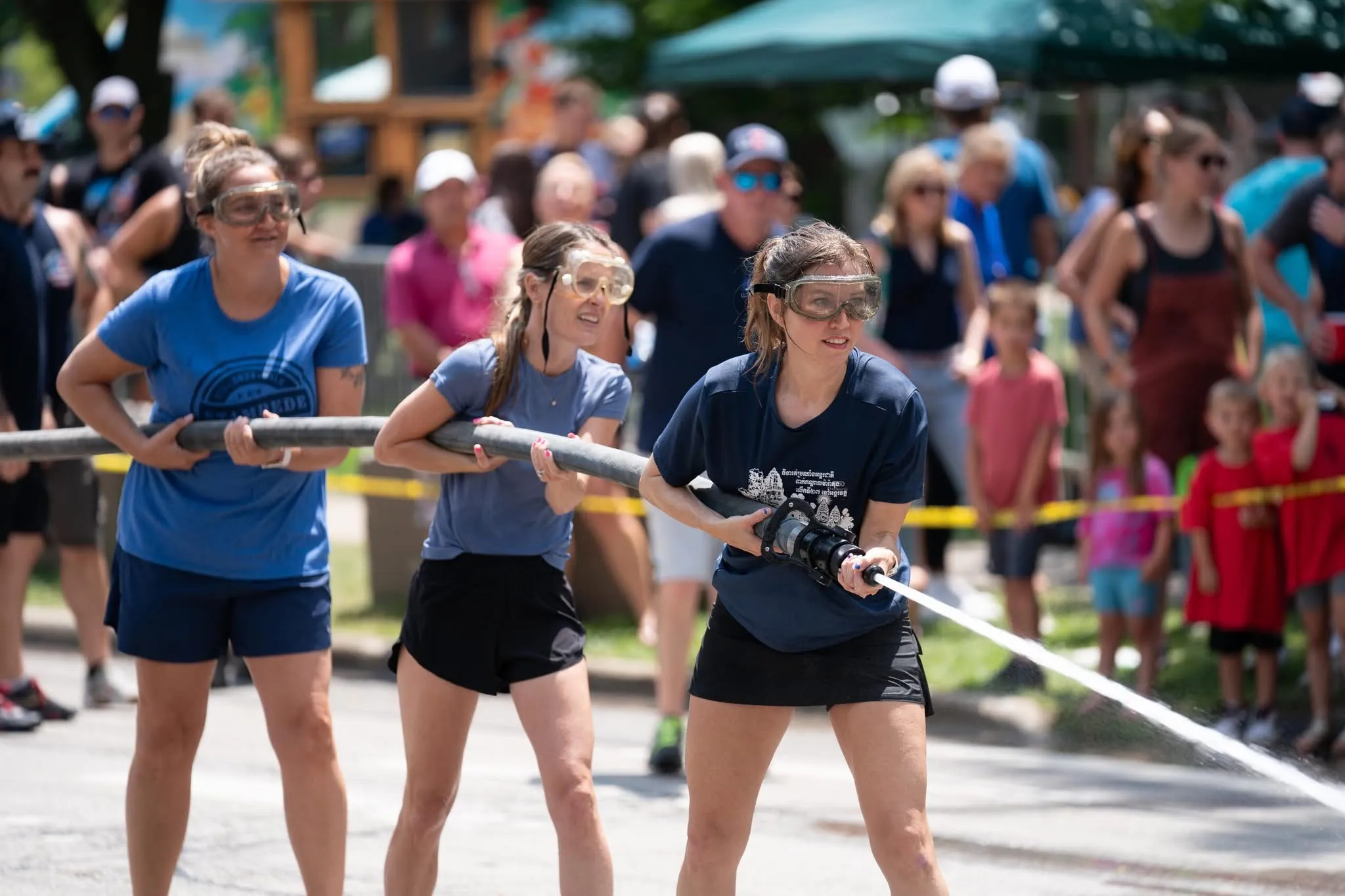 Three women in goggles brace a charged fire hose during the Frontier Days water fights competition, the front woman aiming a jet of water as a crowd of spectators watches from behind the safety line on a sunny day.