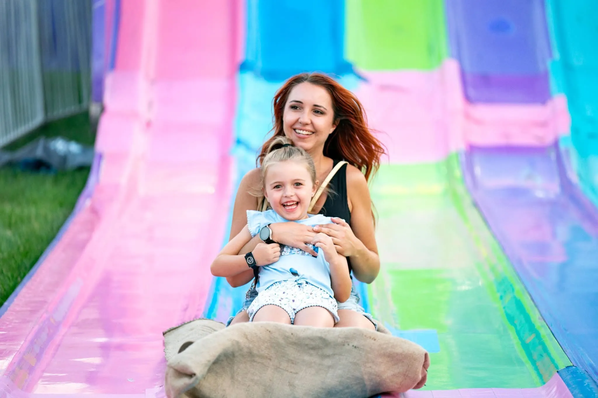 A laughing mother and her young daughter slide down a colorful giant carnival slide together on a burlap sack at Frontier Days, both smiling at the camera on a sunny day.