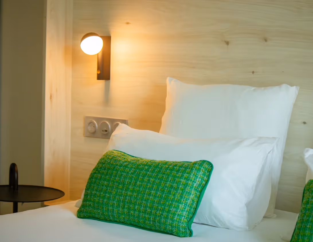 Détail de la tête de lit en bois clair avec coussin vert et applique dorée de la chambre Ysotop à Grenoble.