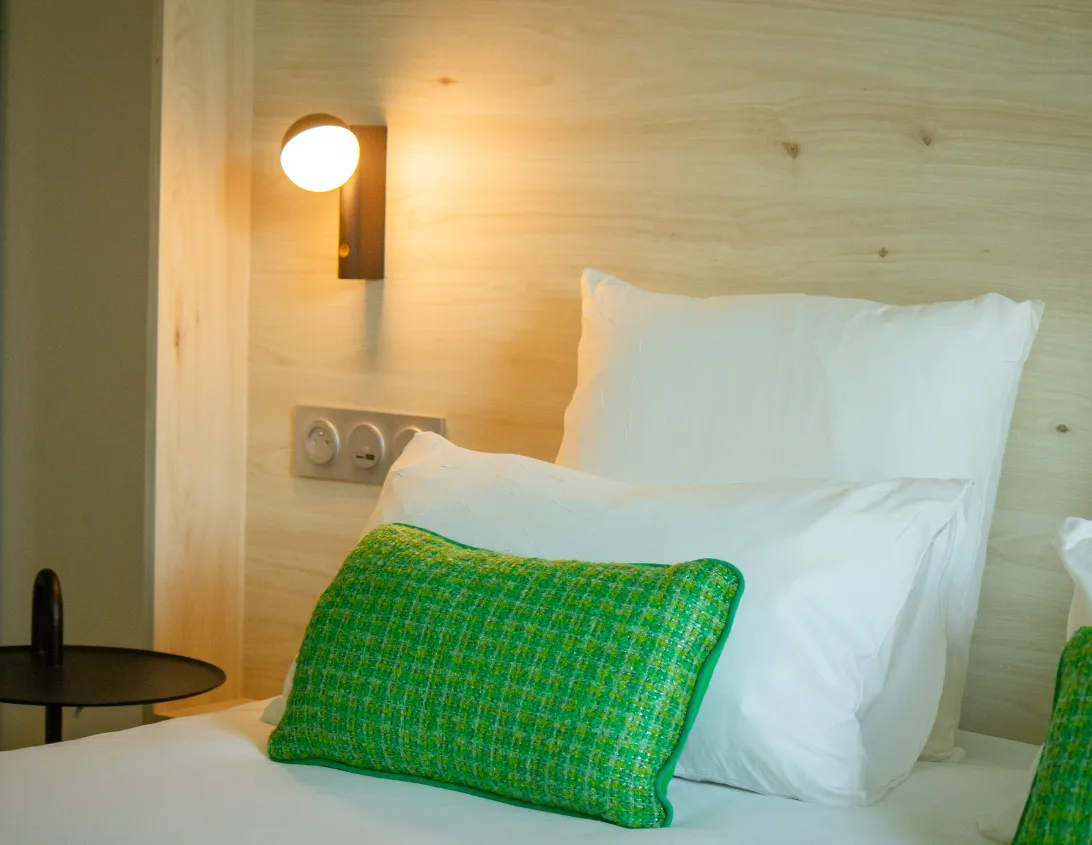 Détail de la tête de lit en bois clair avec coussin vert et applique dorée de la chambre Ysotop à Grenoble.