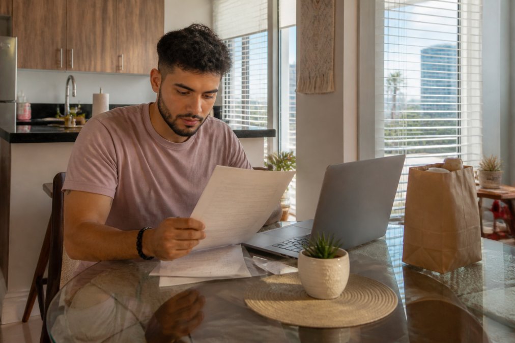 A patient reviewing insurance paperwork at a kitchen table in a Miami apartment, laptop open and a pharmacy bag nearby, warm afternoon light through the blinds
