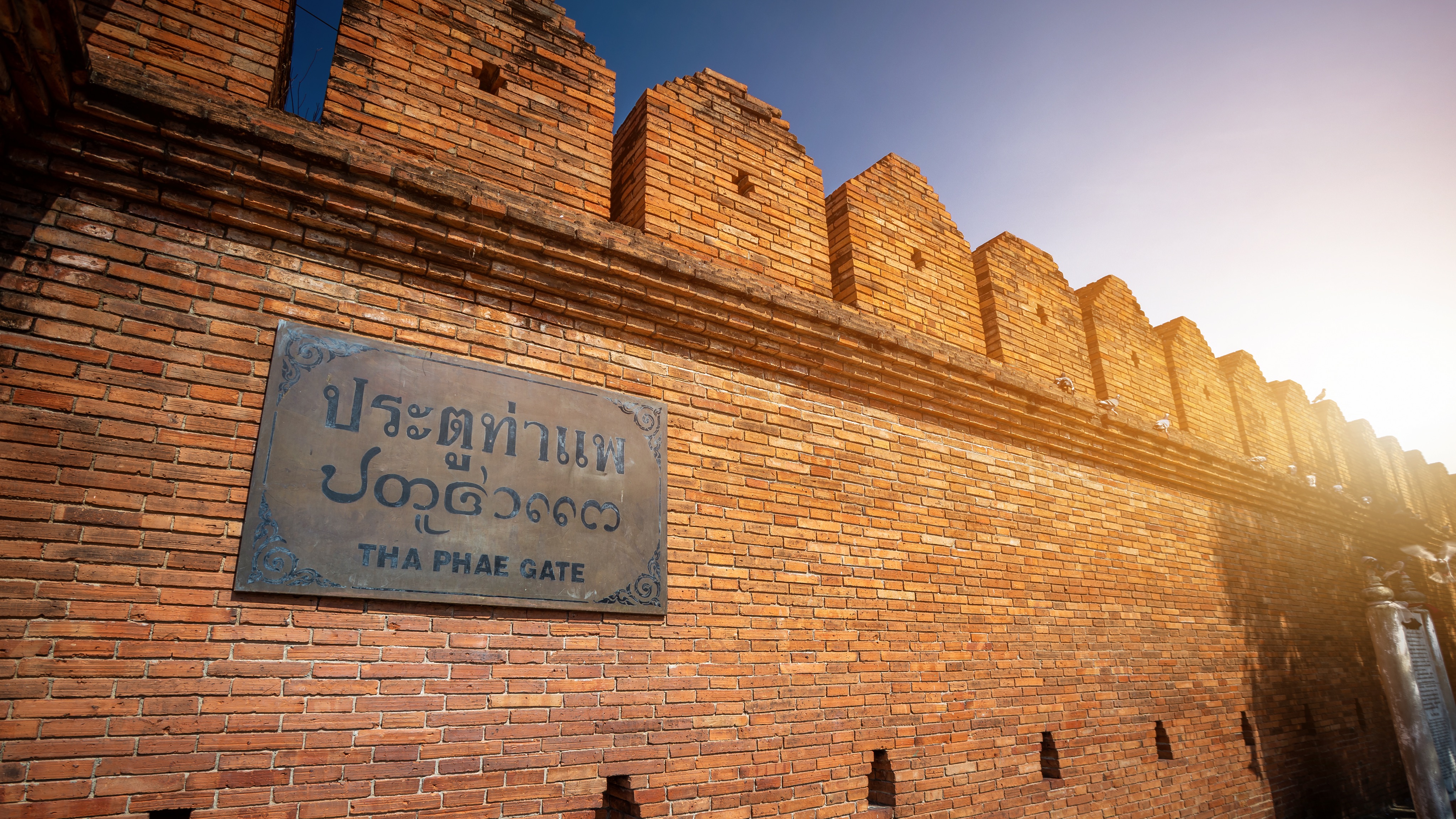 Sunlit red brick wall of Tha Phae Gate in Chiang Mai, Thailand with a metal sign in Thai and English.