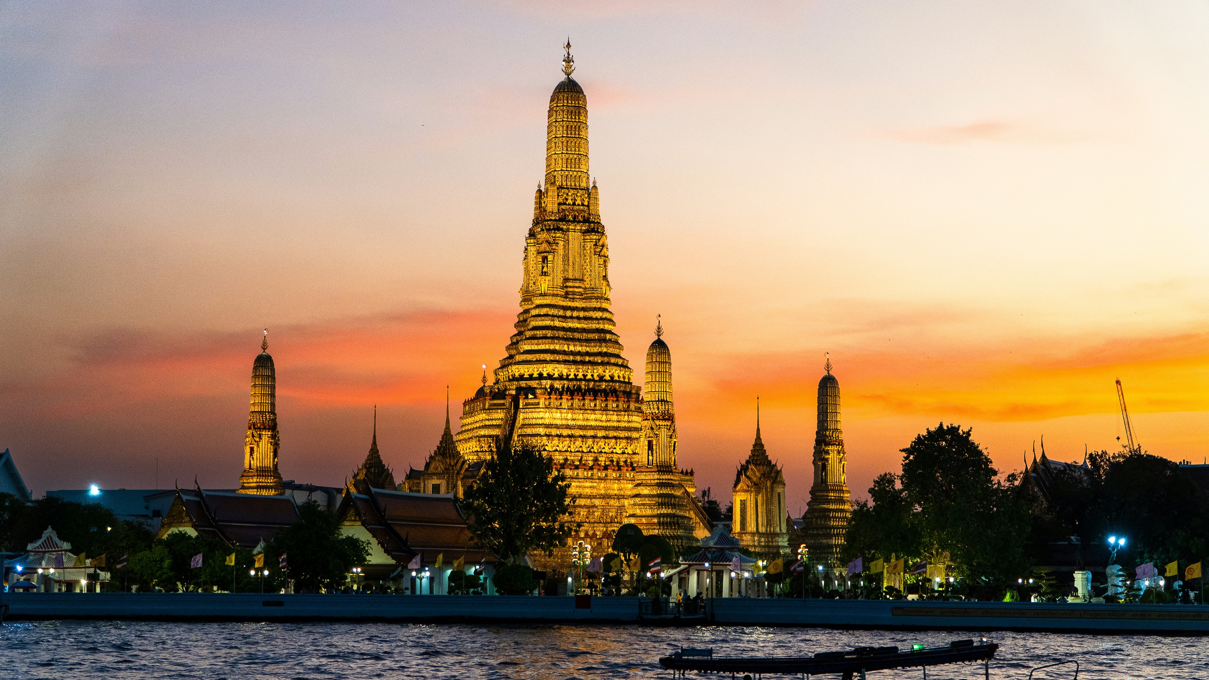 Illuminated Wat Arun temple on the riverbank during sunset with a colorful sky in the background.