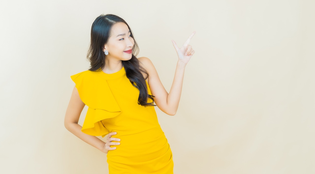 Woman in yellow dress smiling and pointing to her right against a beige background.