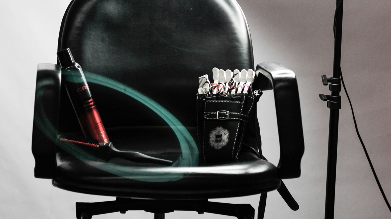 Black salon chair with hair styling products and tools on the seat and a stand light beside it.