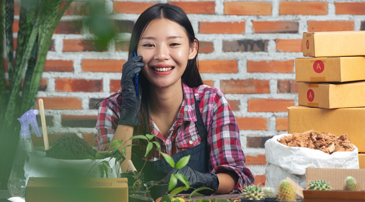 Smiling woman wearing gloves talks on phone near plants and stacked cardboard boxes on table.