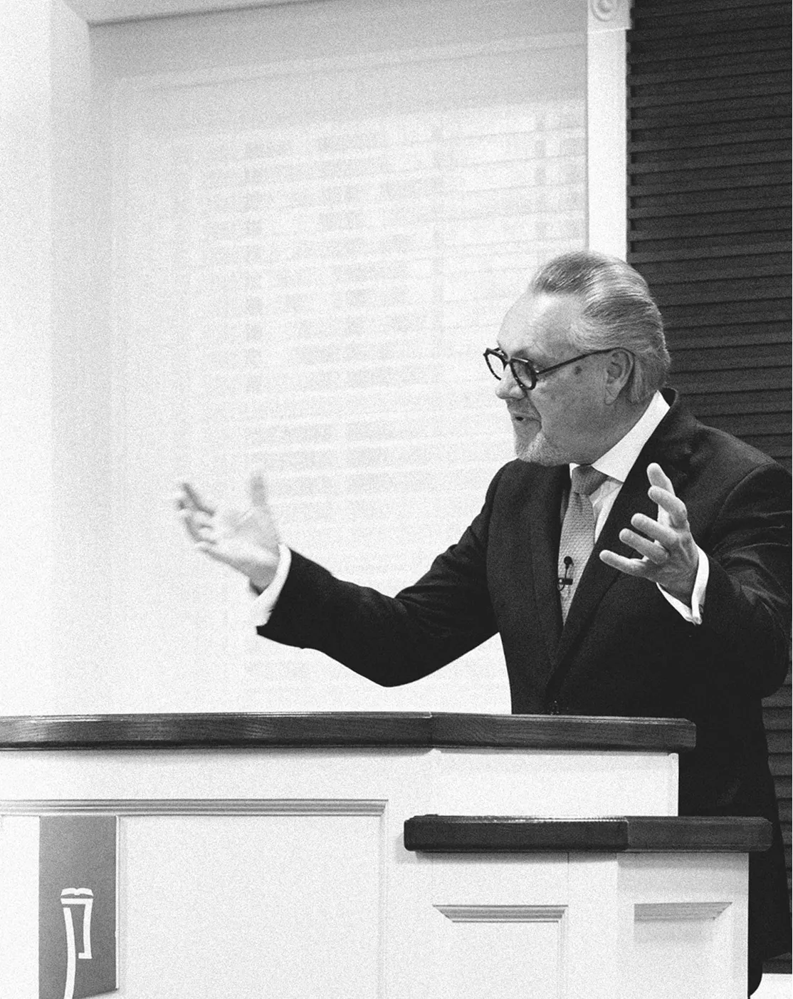 Man in suit and glasses speaking passionately behind a white podium with gesturing hands.