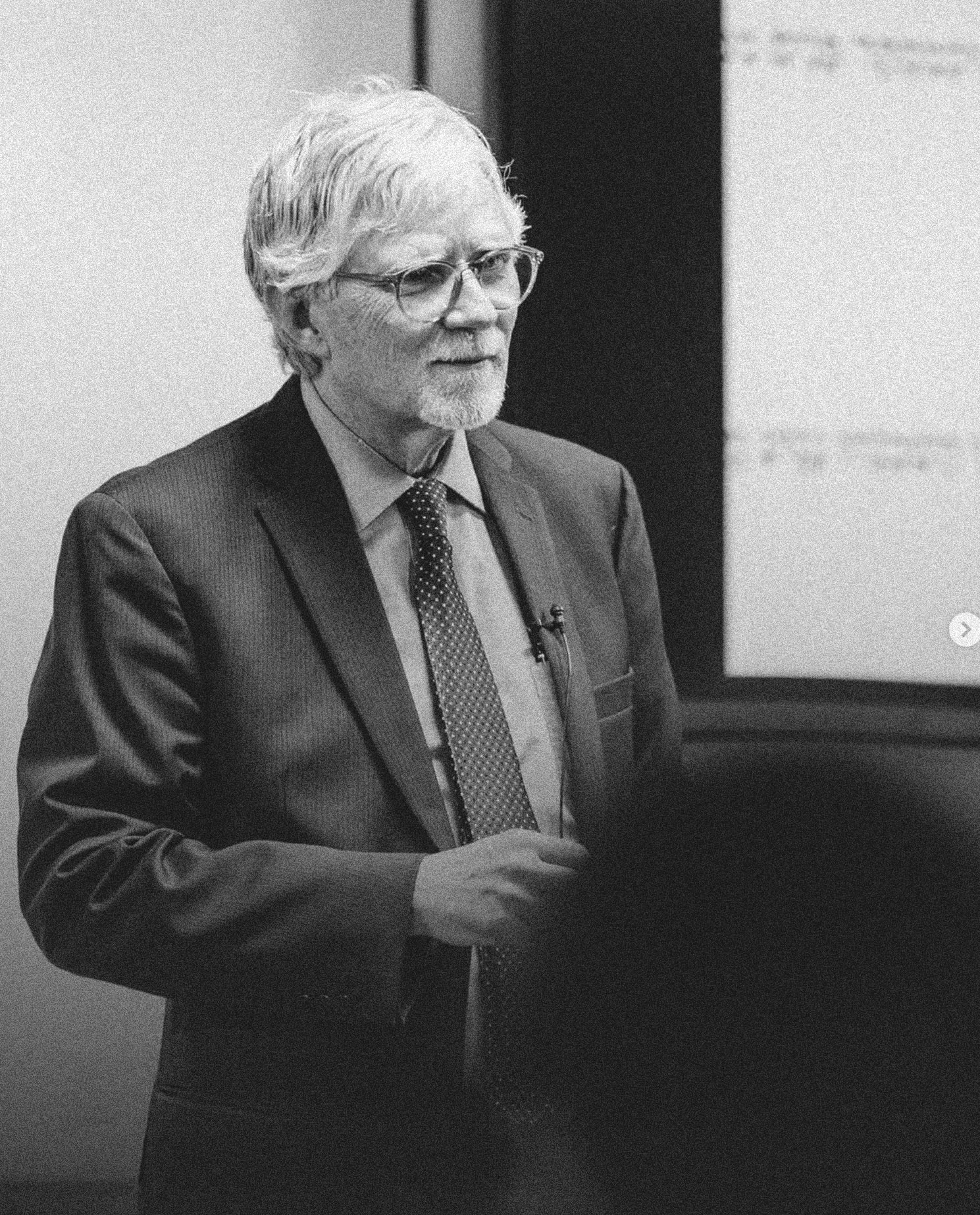 Older man with white hair and glasses wearing a suit and tie, speaking or presenting in front of a screen.