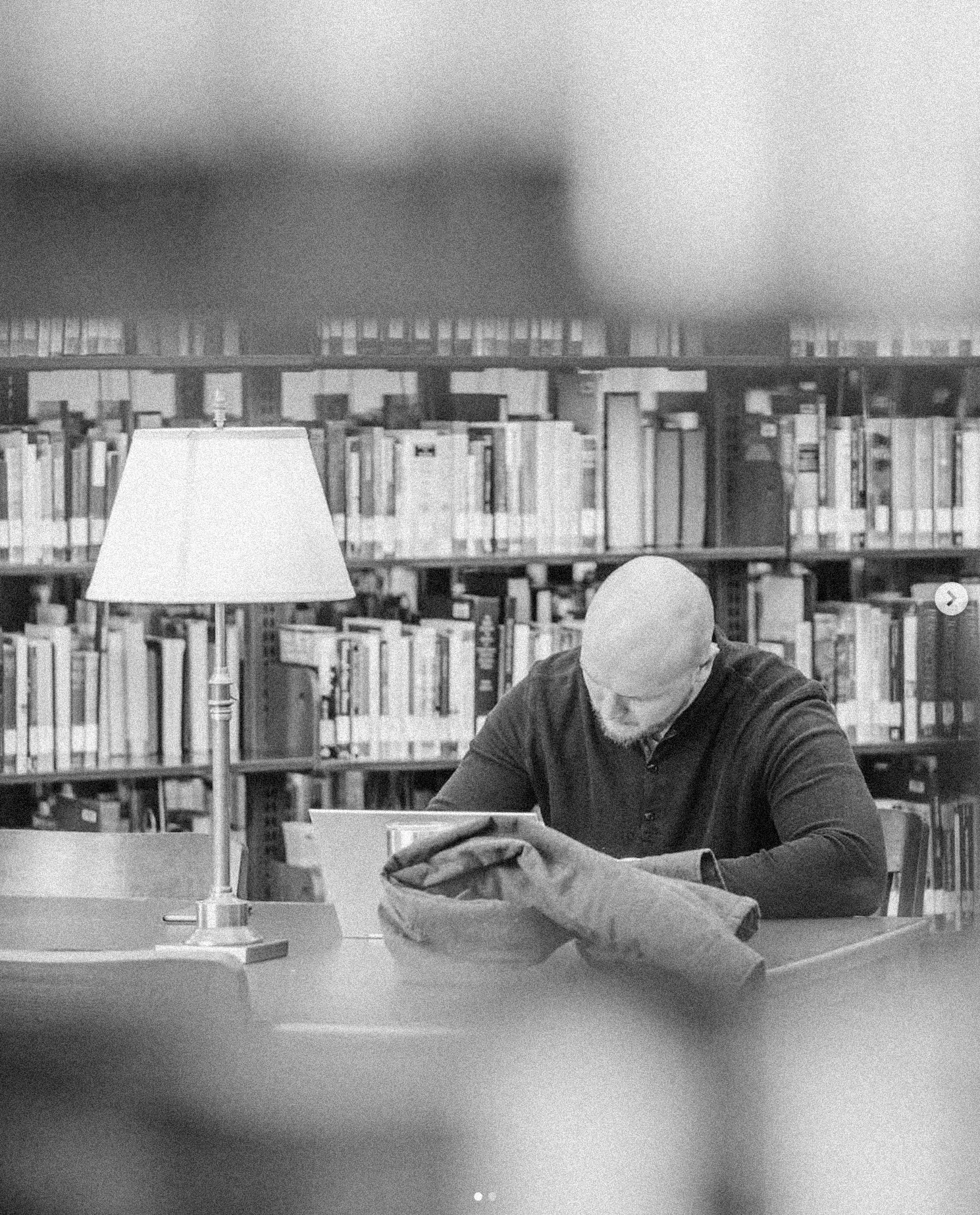 Man reading a book at a library table with a lamp and bookshelves in the background.