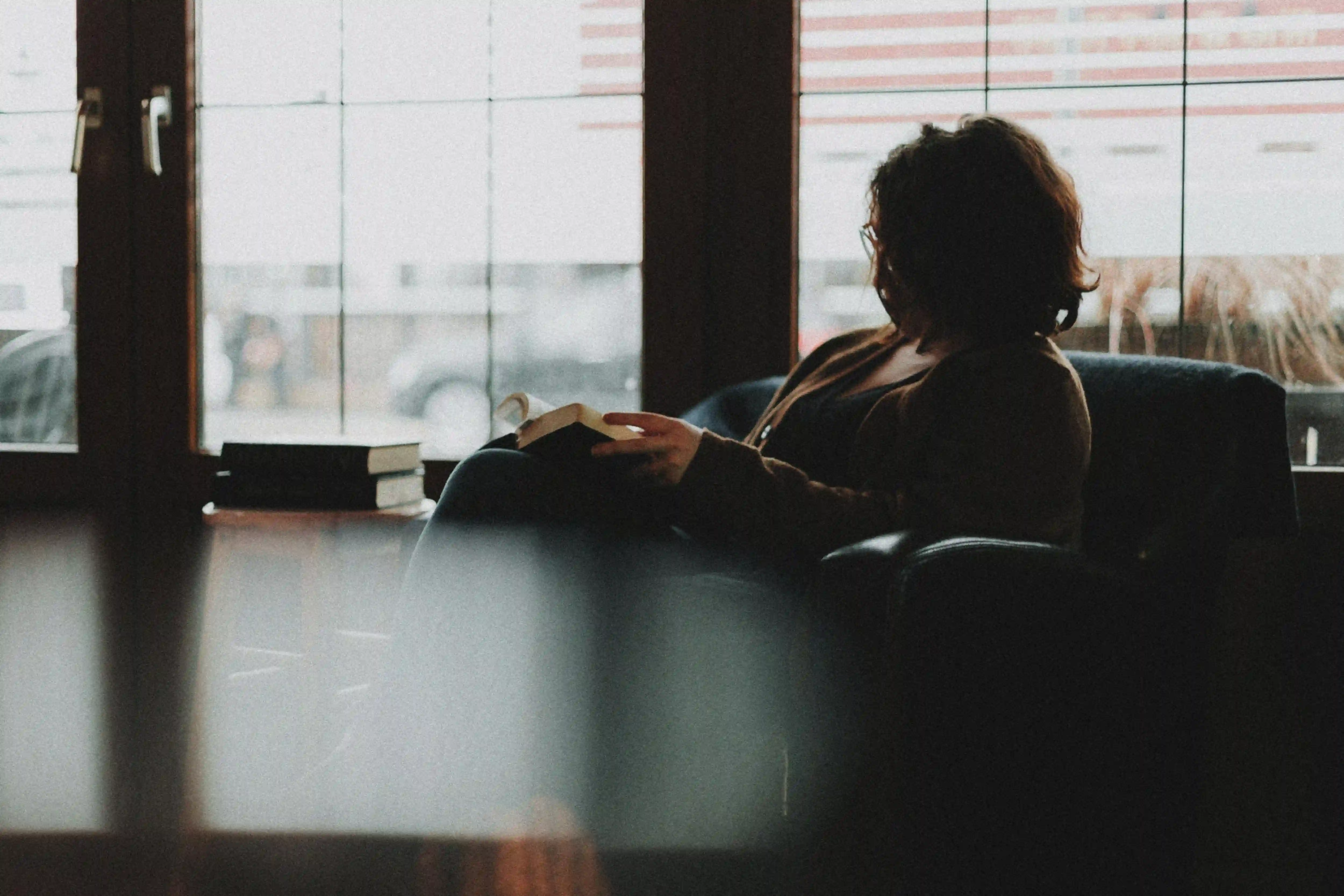Person with curly hair sitting in an armchair by a large window, reading a book.