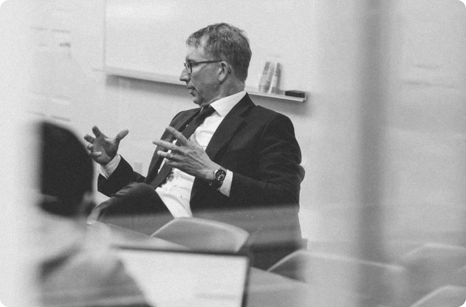 Black and white photo of a man in a suit and glasses speaking and gesturing with his hands in a meeting room.