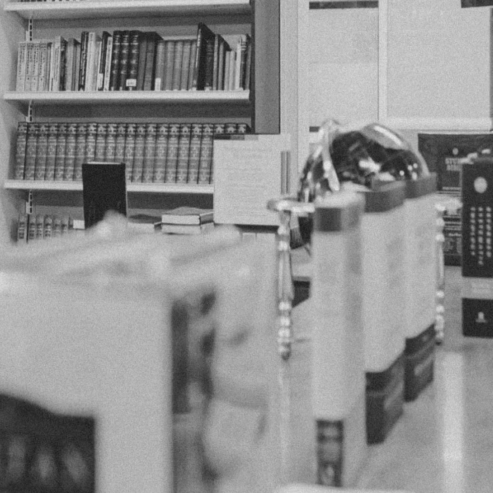 Black and white photo of a bookshelf filled with books and a table with several stacked books and items in the foreground.