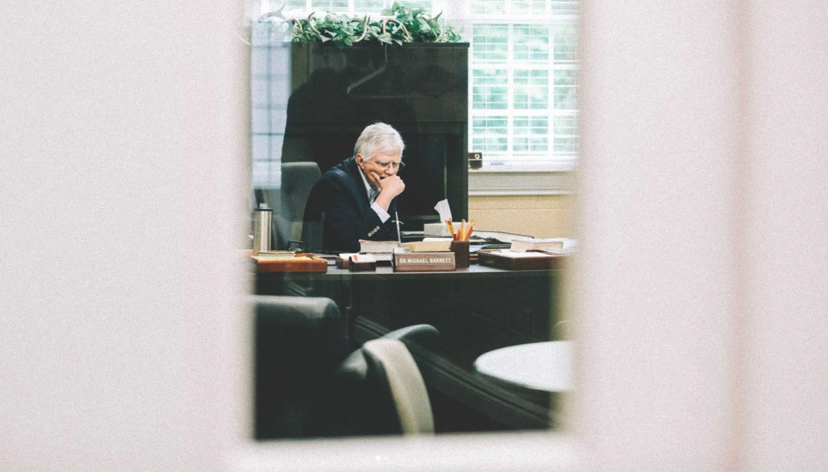 Older man with gray hair in a suit sitting at a cluttered desk reading papers in a bright office.