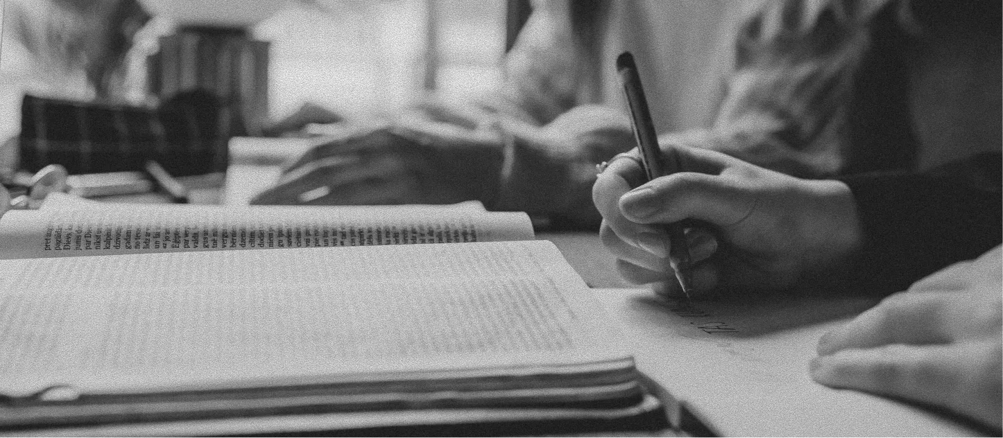 Close-up of hands writing with a pen on paper next to open books in a scholarly setting.