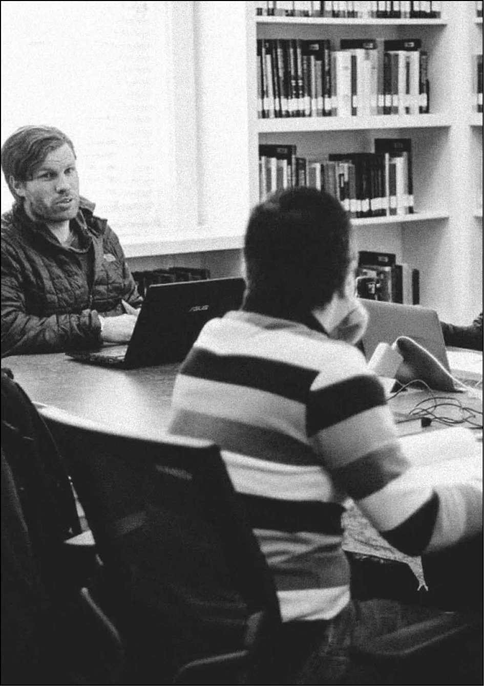Two men sitting at a table in a library or study room with laptops and books, engaged in conversation.