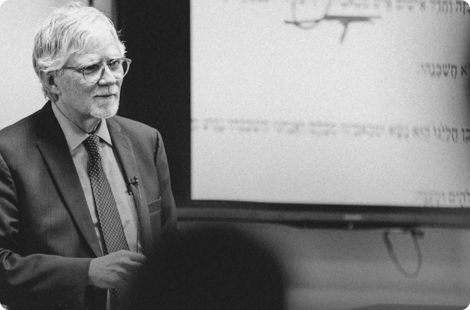 Elderly man with white hair and glasses wearing a suit and tie, speaking in front of a blurred screen with text.