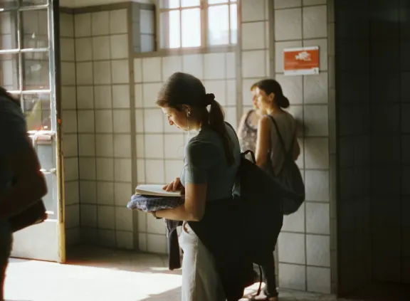 Young woman reading a book in a tiled hallway with sunlight coming through the door.