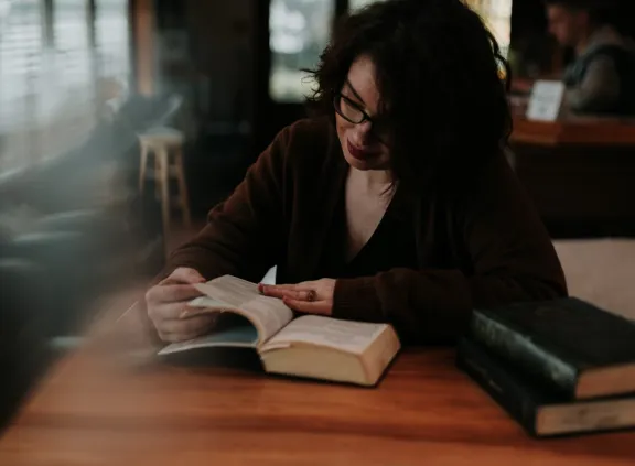 Woman with glasses reading a book at a wooden table in a dimly lit room.