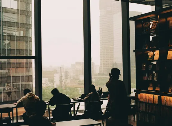 Silhouettes of people seated and standing near large windows overlooking city buildings, inside a library or study area.