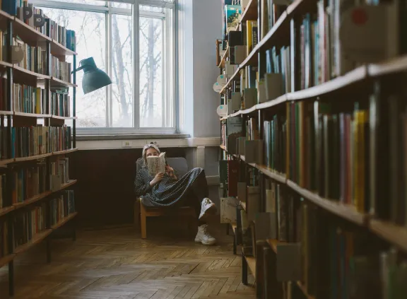 Person sitting in a library chair near a window, reading a book surrounded by bookshelves.