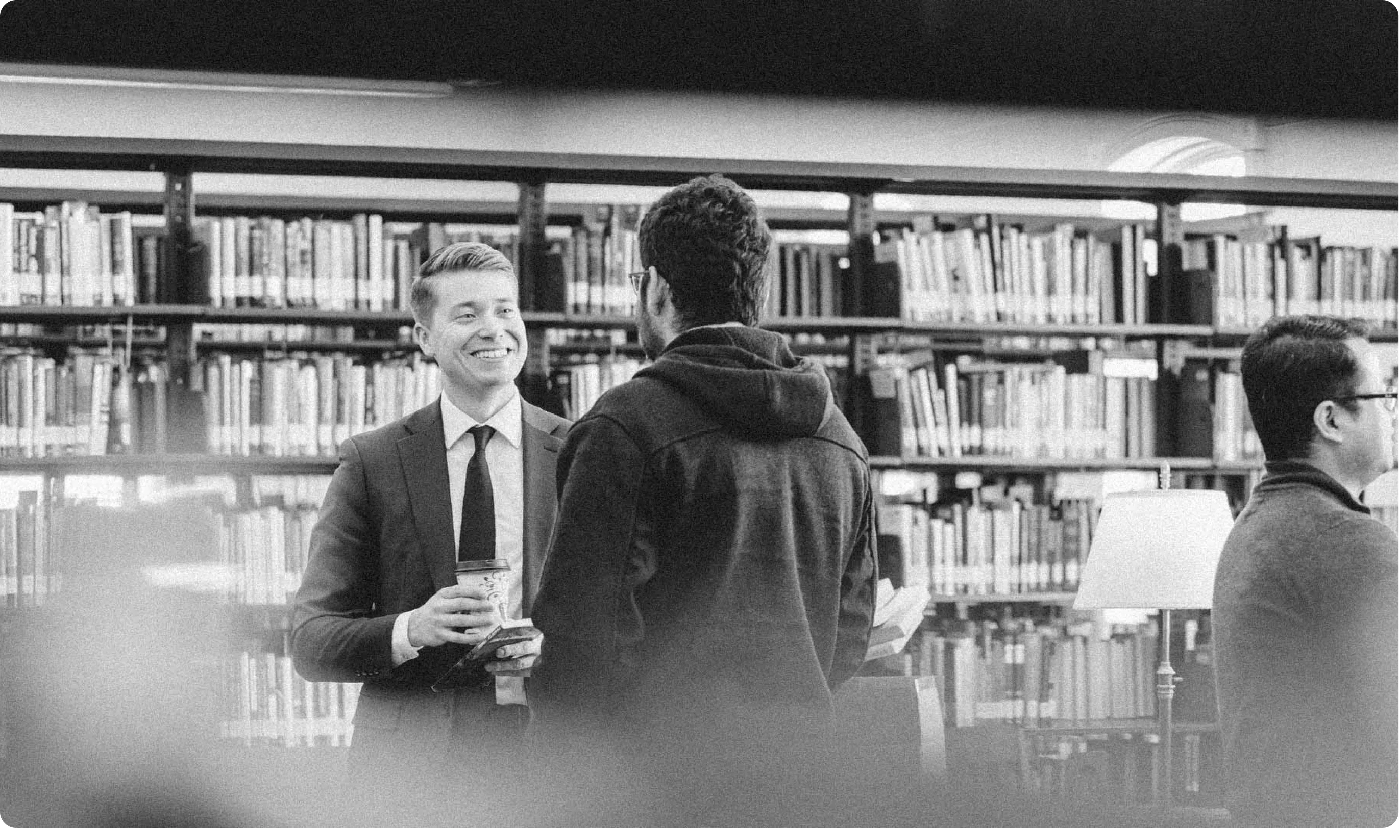 Two men having a conversation in a library with bookshelves in the background.