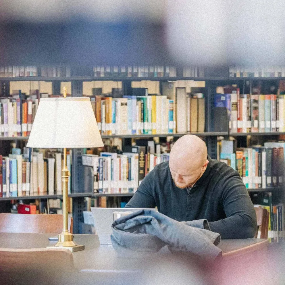 Man with shaved head studying at a library table with a lamp and bookshelves in the background.