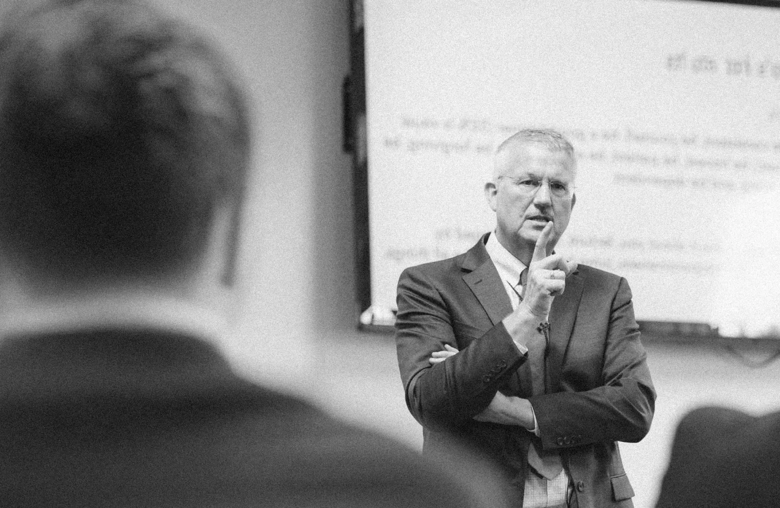 Man in suit speaking thoughtfully with finger near mouth in front of a blurred screen in a classroom.