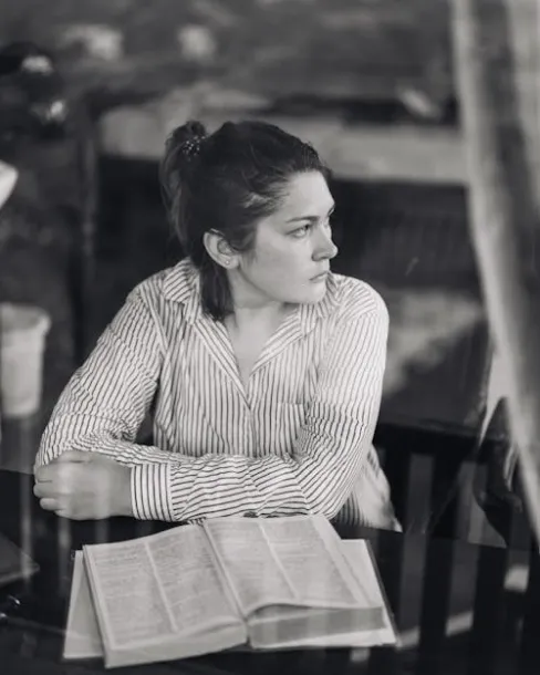 Woman in a striped shirt sitting at a table with an open book, looking thoughtfully to the side.