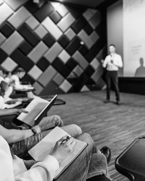 Black and white image of students taking notes while a speaker presents in front of a patterned wall.
