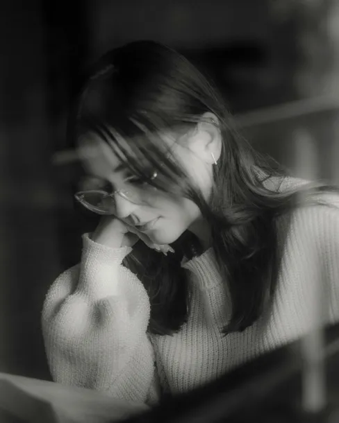 Black and white photo of a woman wearing glasses and a sweater, reading with her head resting on her hand.