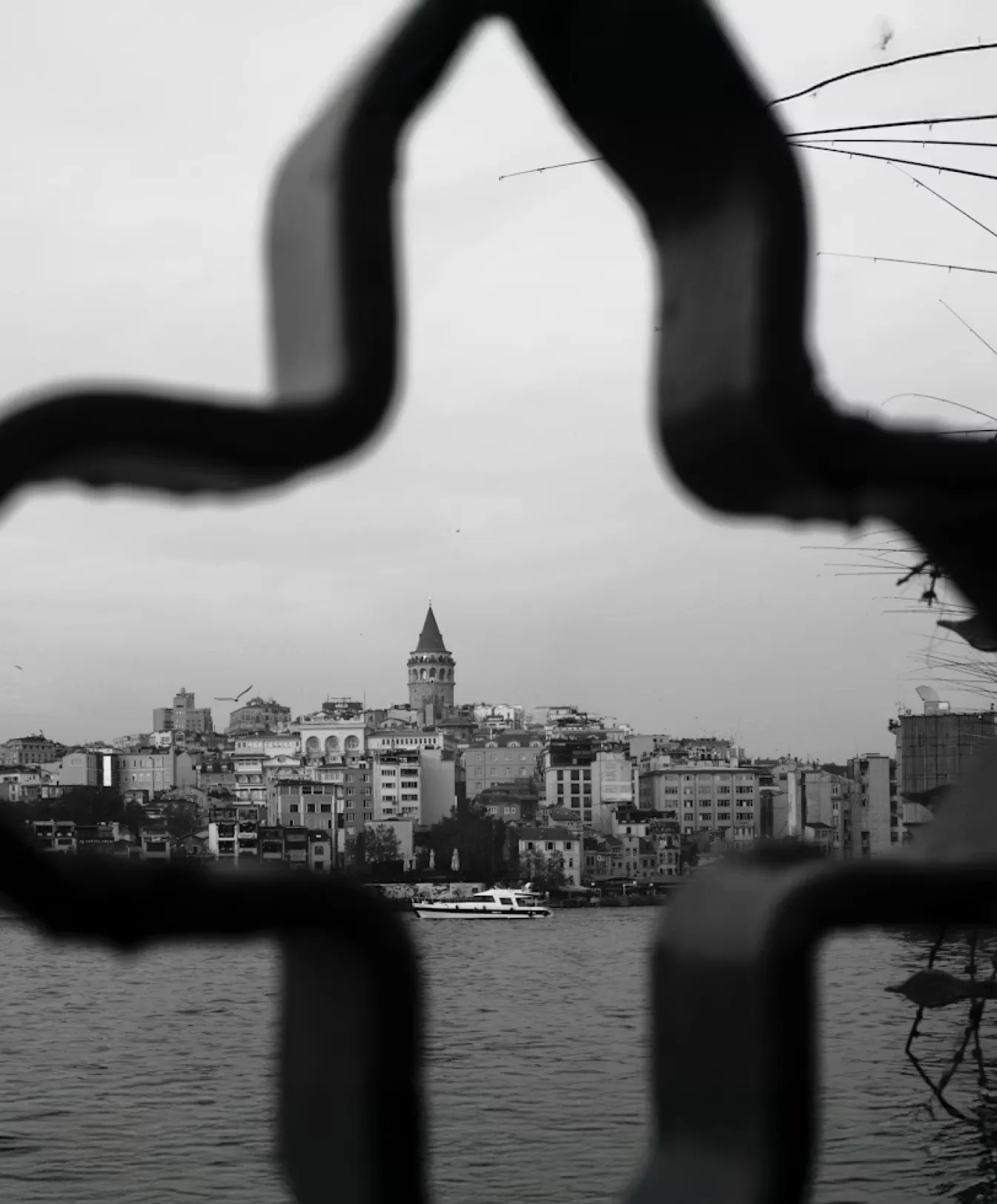 Black and white cityscape of buildings and Galata Tower framed by a blurred star-shaped metal structure over water.