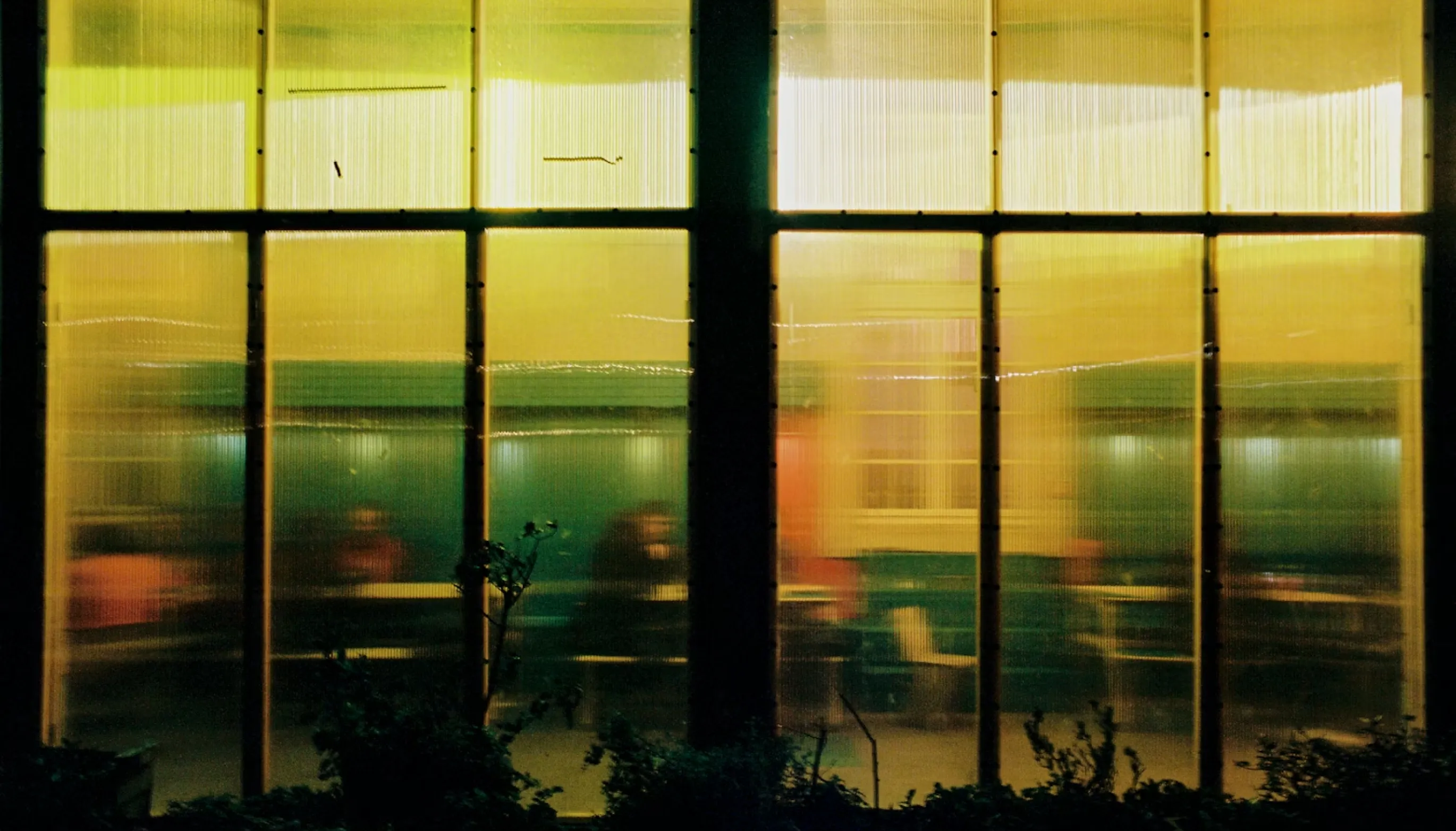 Blurred silhouettes of people sitting inside a dimly lit room seen through frosted glass windows at night.