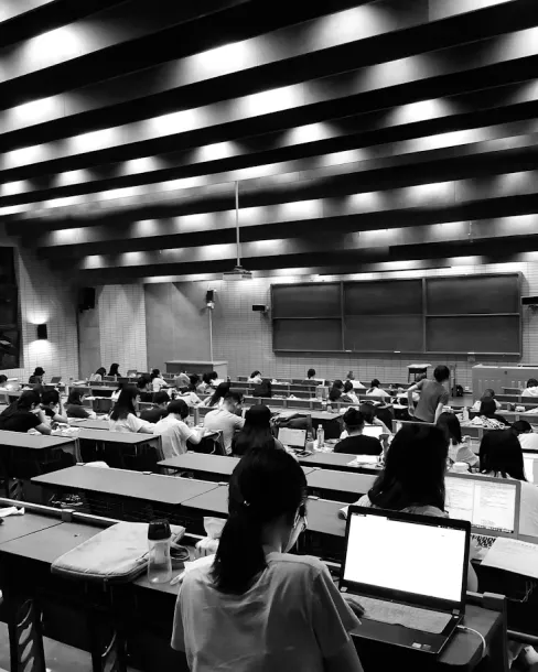 Black and white photo of a large lecture hall filled with students studying at desks with laptops and books.