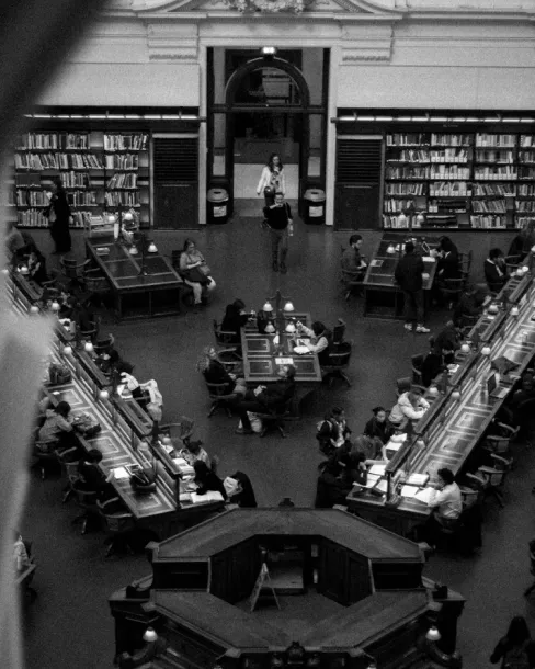 Black and white photo of a library reading room with people seated at desks reading and studying.