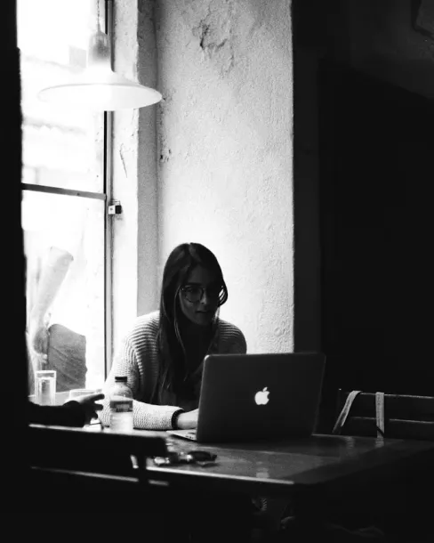 Woman wearing glasses working on a MacBook laptop at a wooden table near a window.