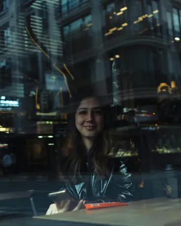 Young woman smiling inside cafe, reflection of city lights and buildings on window glass.