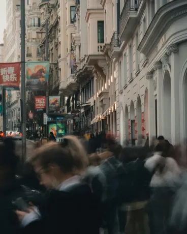 Blurred crowd of people walking on a city street lined with tall buildings and signs.