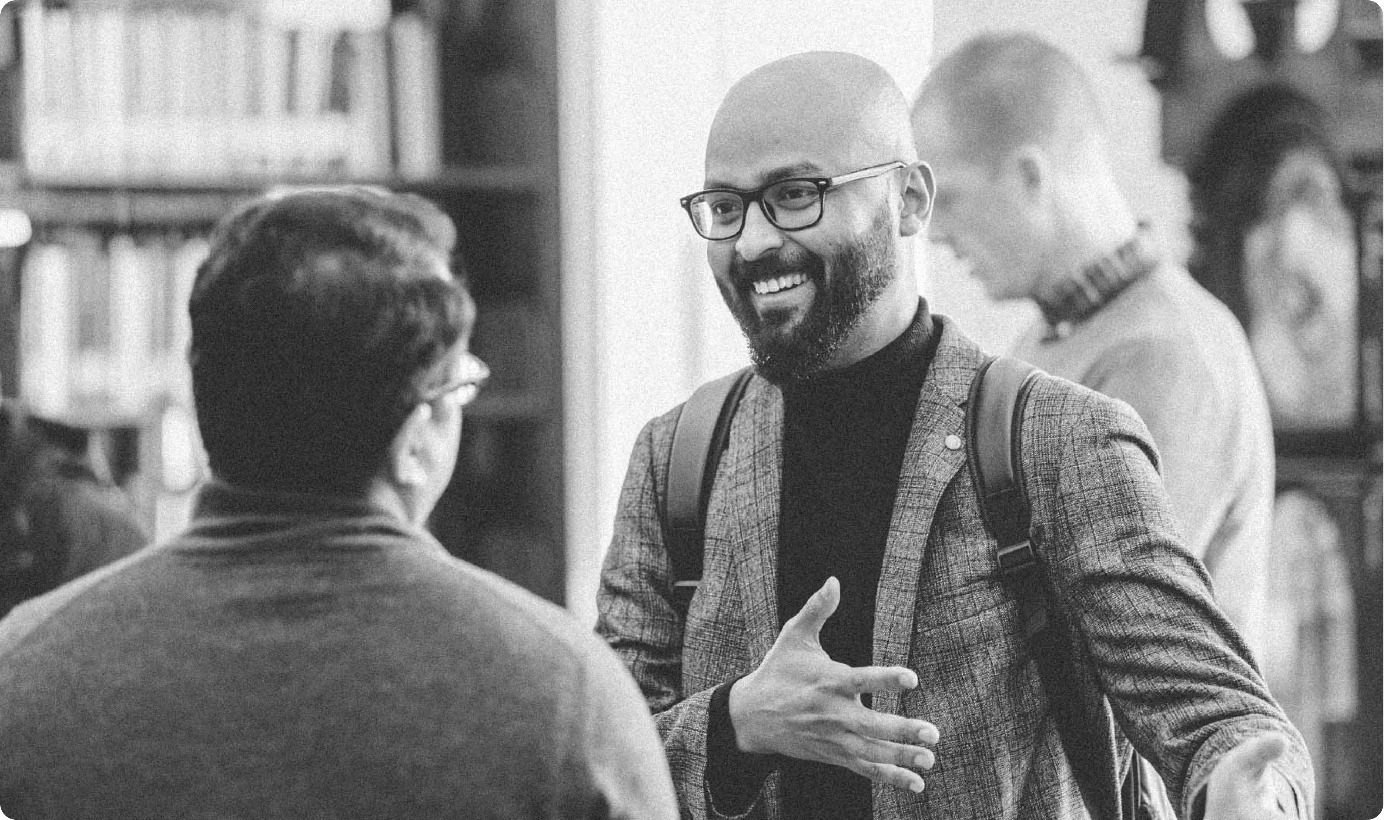Two men in conversation; one smiling, wearing glasses and a plaid jacket with a backpack.