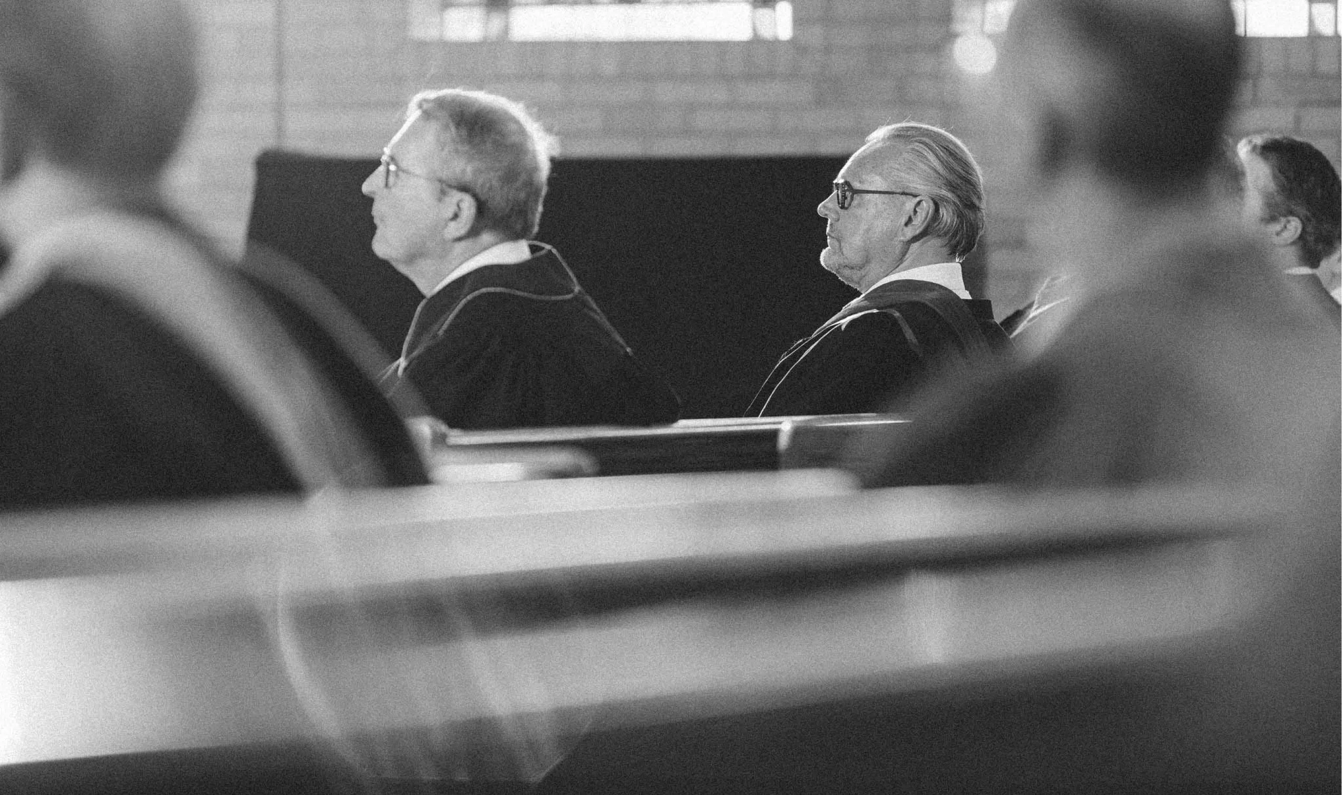 Black and white photo of men in academic gowns sitting in pews or benches inside a building.