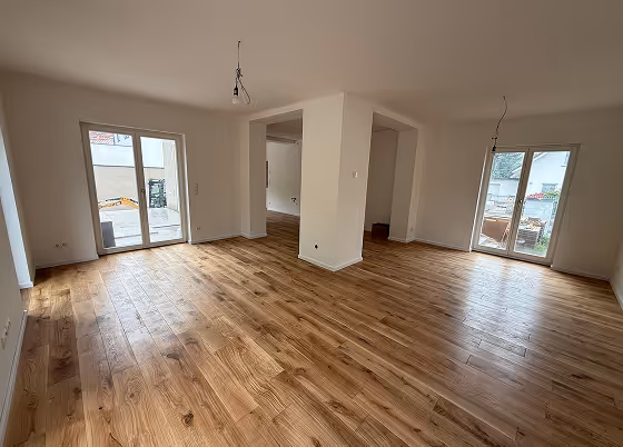Großes, leeres Zimmer mit hellem Holzboden, weißen Wänden, zwei Glastüren zu einer Terrasse und minimaler Beleuchtung.