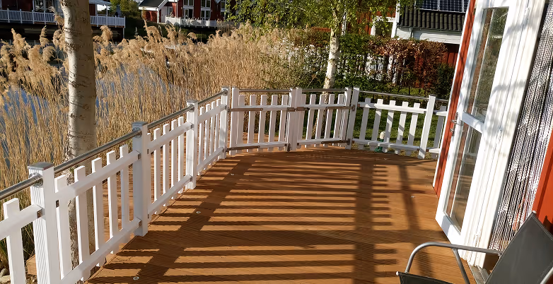 Holzterrasse mit weißem Zaun und Gartenstühlen neben einem Haus, mit Bäumen und hohen Gräsern im Hintergrund.