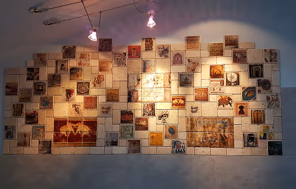 Beleuchtete Wand mit einer Installation aus zahlreichen kleinen quadratischen Kacheln, die verschiedene Zeichnungen und Muster zeigen.