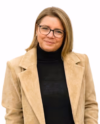 Frau mit blonden Haaren, Brille und beiger Jacke über schwarzem Pullover lächelnd vor weißem Hintergrund.