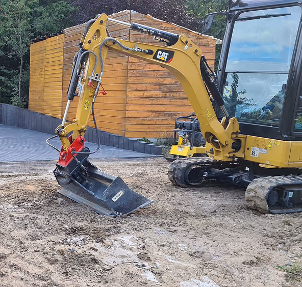 Gelber Caterpillar-Minibagger auf einer Baustelle mit freigelegtem Boden und einem Holzanbau im Hintergrund.