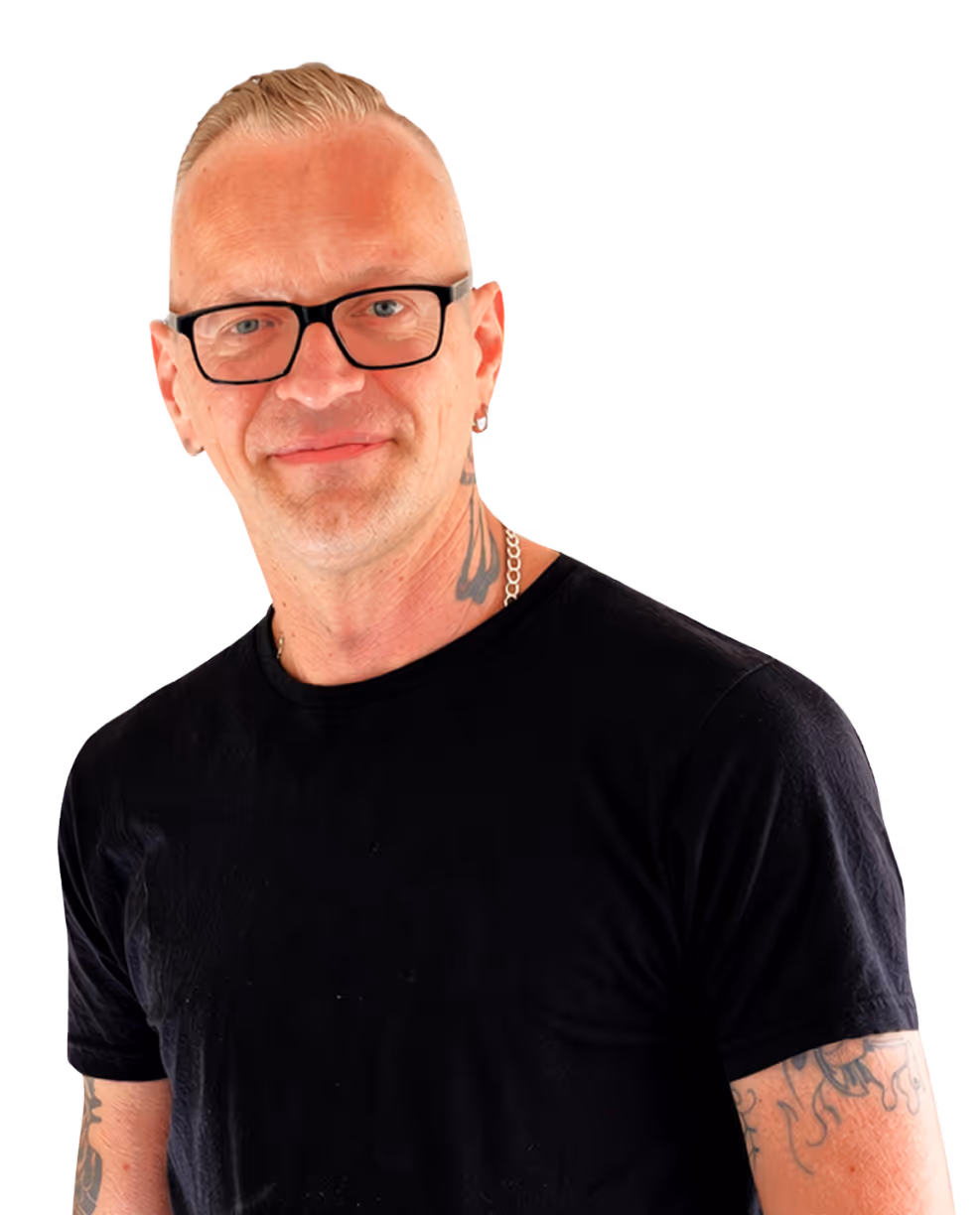 Mann mit kurzen blonden Haaren, schwarzer Brille, schwarzen T-Shirt, Tattoos am Hals und Arm, lächelnd vor schwarzem Hintergrund.