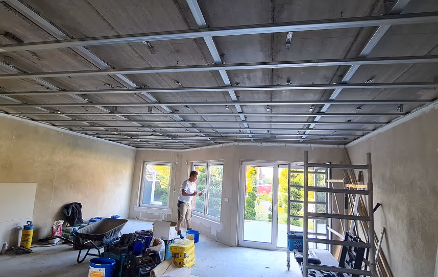 Person bei der Renovierung eines Raums mit sichtbaren Metallträgern an der Decke, einer Schubkarre, Werkzeugen und Baustoffen im Inneren.